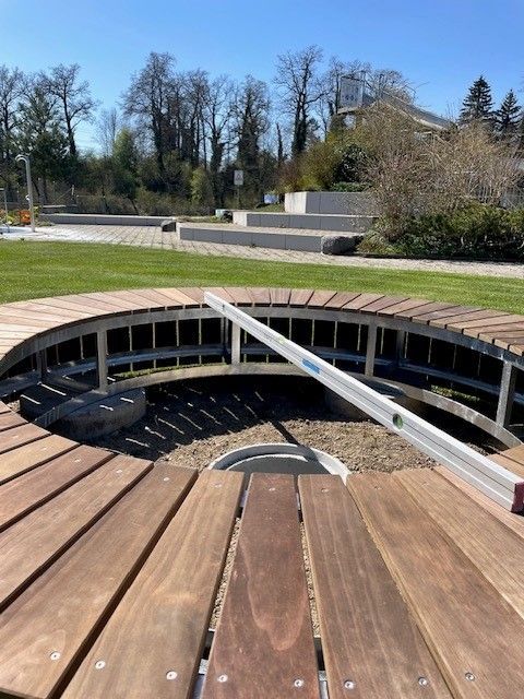 Eine runde Holzterrasse umgibt eine versenkte Feuerstelle in einem Park, im Hintergrund sind Gras und Bäume zu sehen.