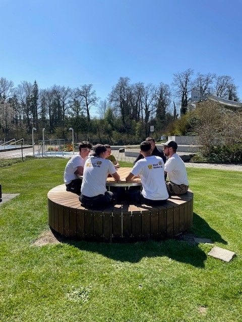 Vier Personen sitzen um einen runden Holztisch im Freien auf einer sonnigen Rasenfläche.