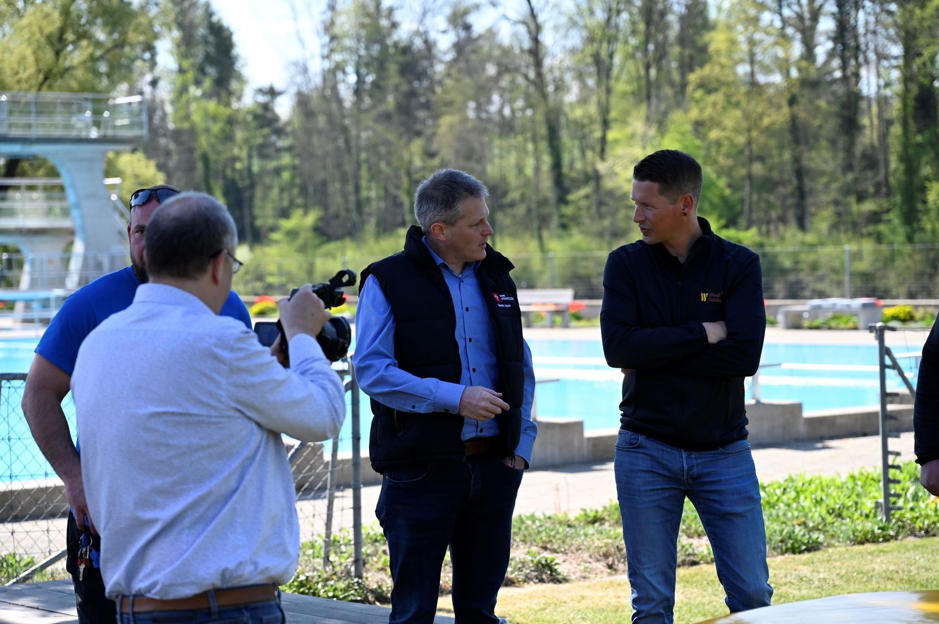 Drei Männer unterhalten sich im Freien in der Nähe eines Pools; im Hintergrund sind Bäume und ein weißes Gebäude zu sehen.