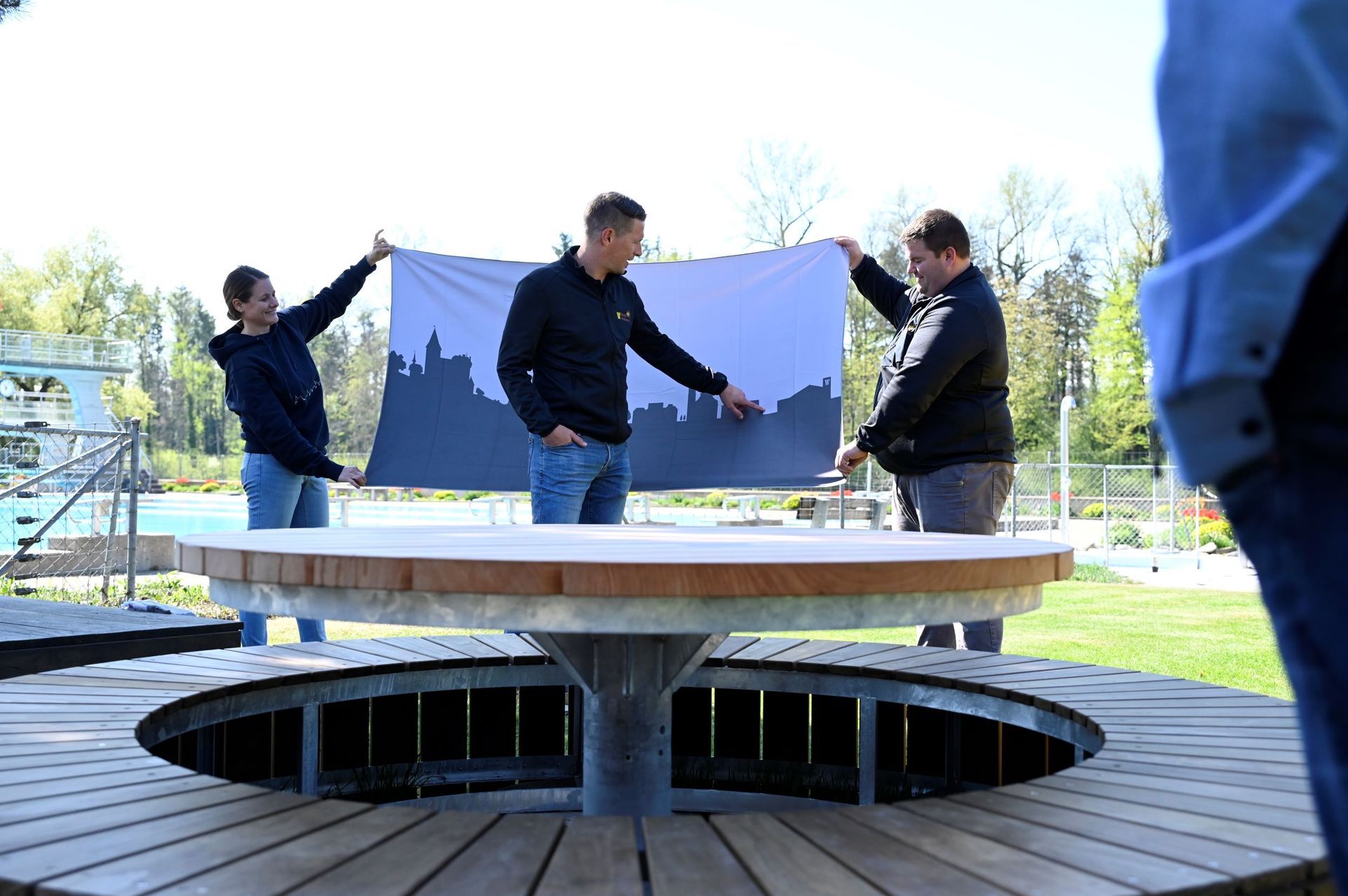 Drei Personen halten ein großes Stoffbanner um einen runden Tisch im Freien auf einer Holzterrasse.
