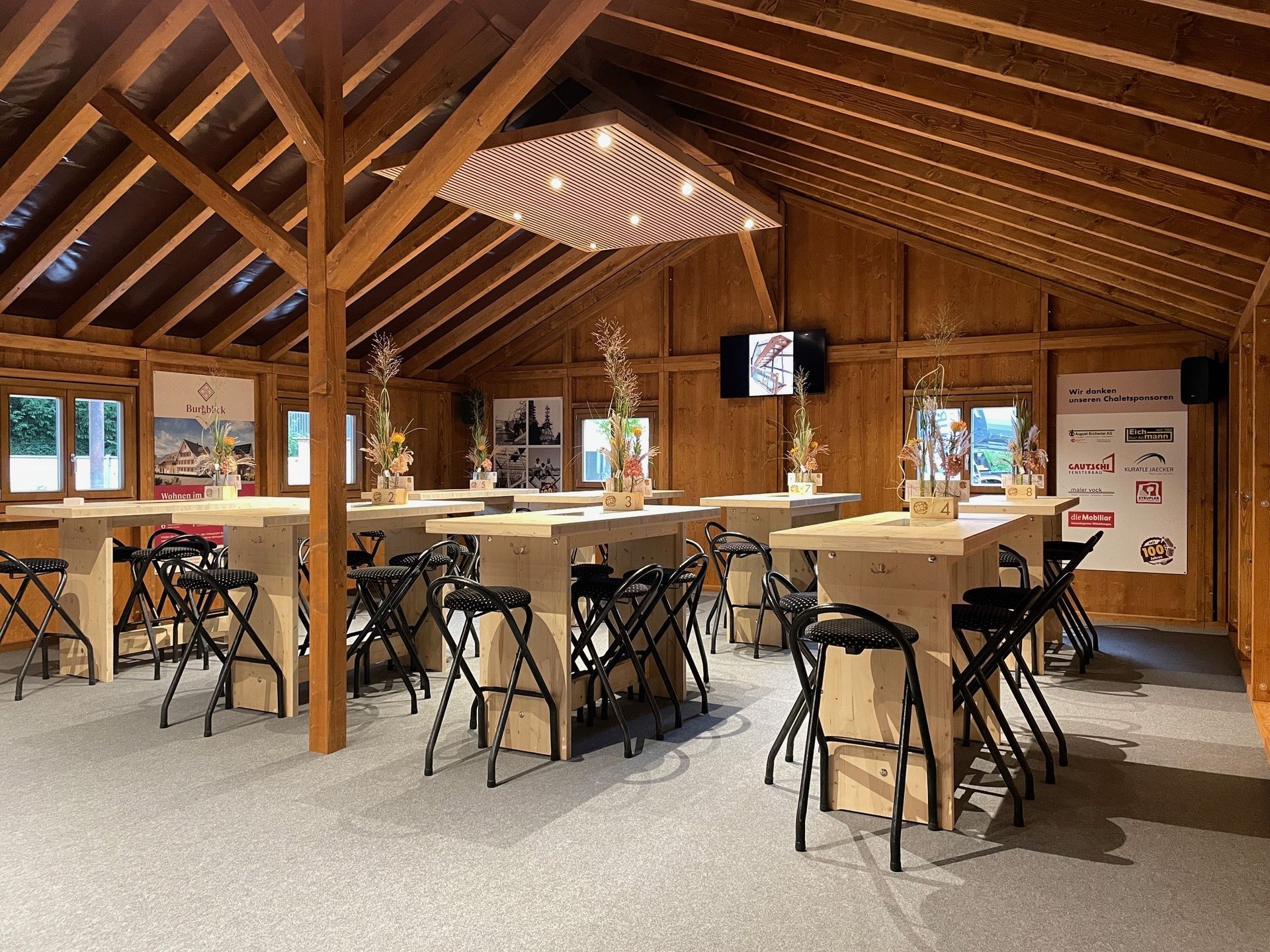 Rustikaler Speisesaal aus Holz mit Picknicktischen und Fernseher, der durch Fenster mit Tageslicht erhellt wird.
