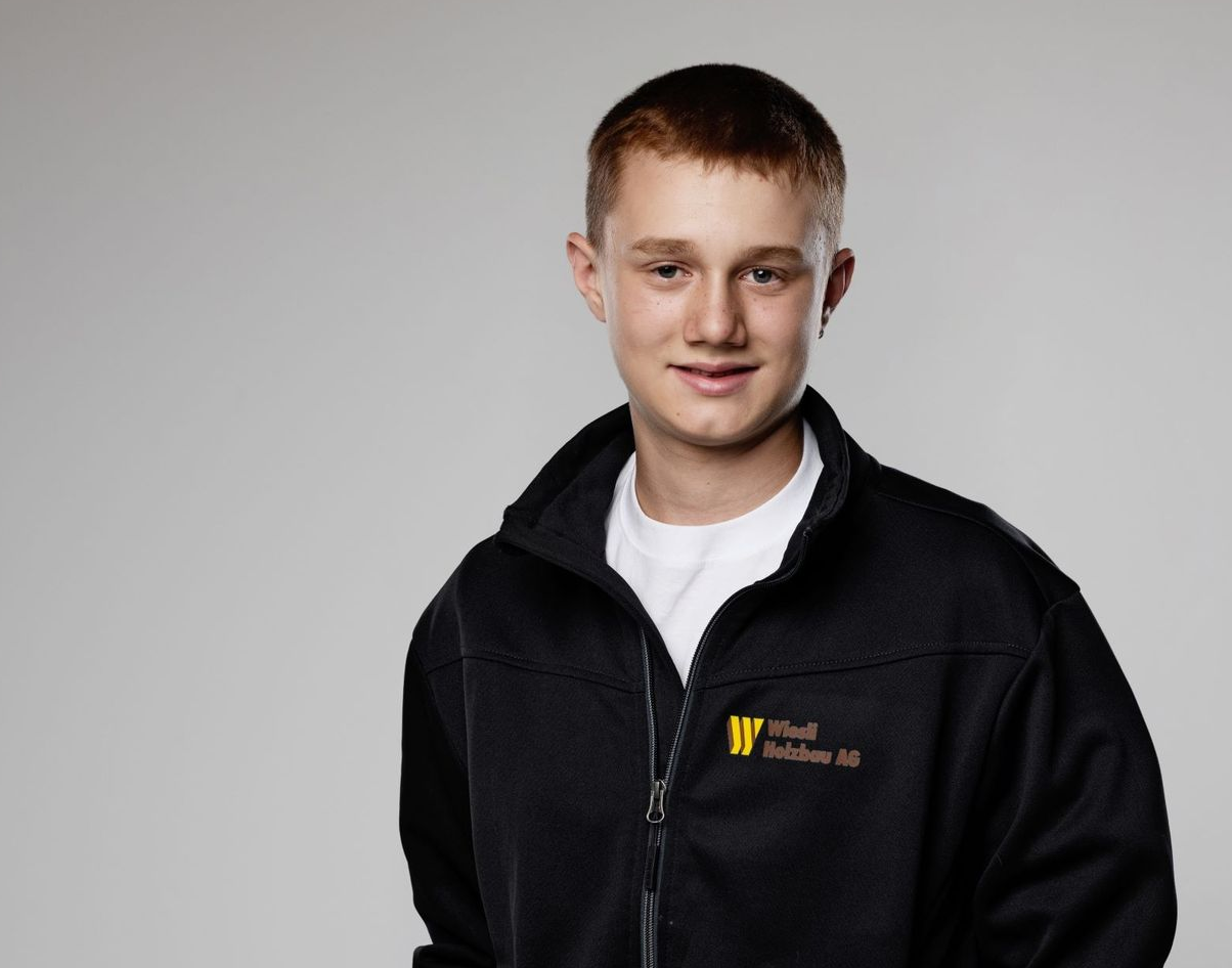 Lächelnde Person in einer schwarzen Reißverschlussjacke über einem weißen Hemd vor grauem Hintergrund
