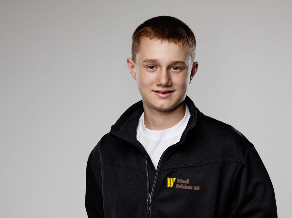 Lächelnde Person in einer schwarzen Reißverschlussjacke über einem weißen Hemd vor grauem Hintergrund