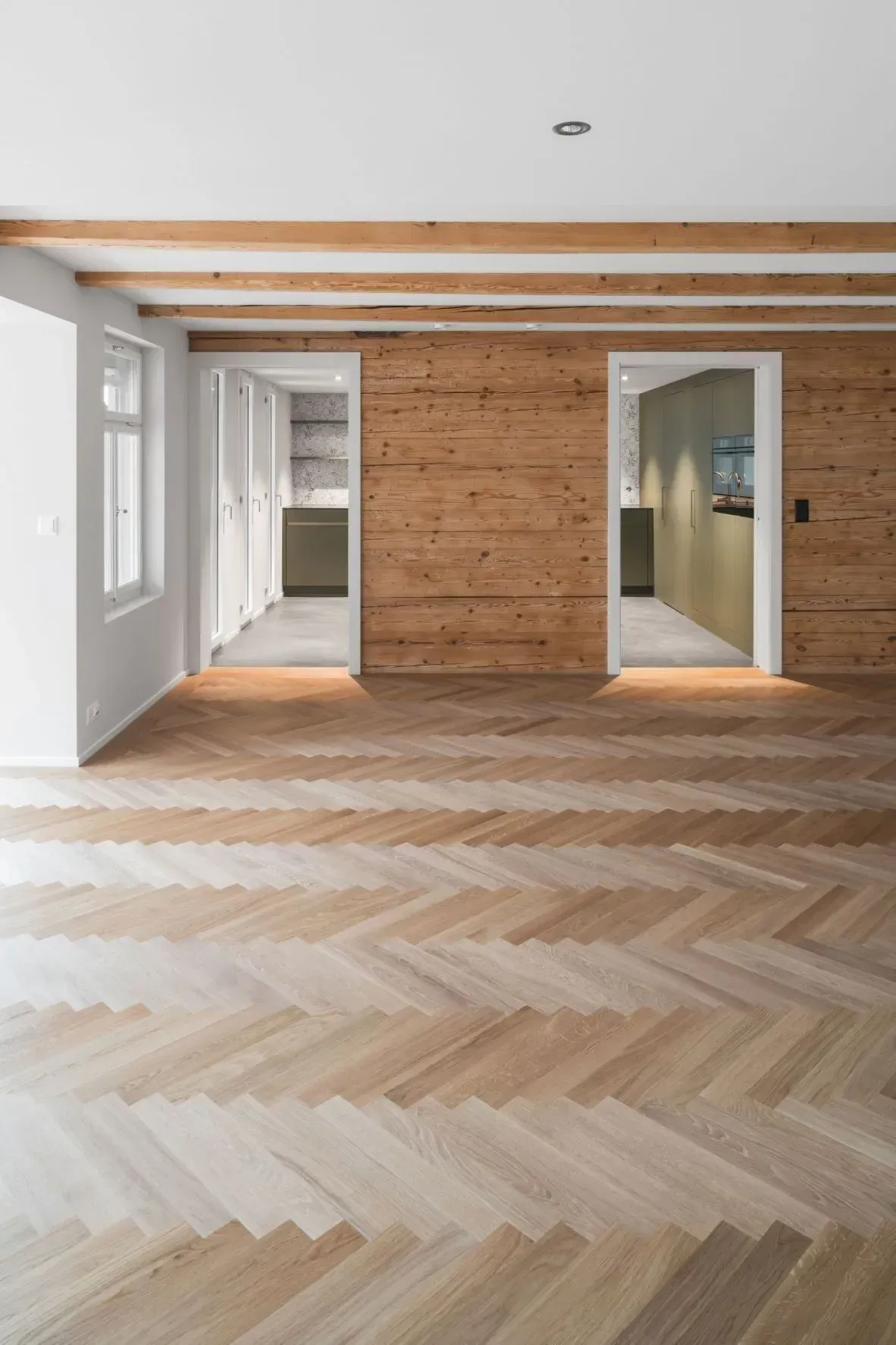 Leeres, modernes Zimmer mit Fischgrätparkett, sichtbaren Deckenbalken und zwei offenen Türen.