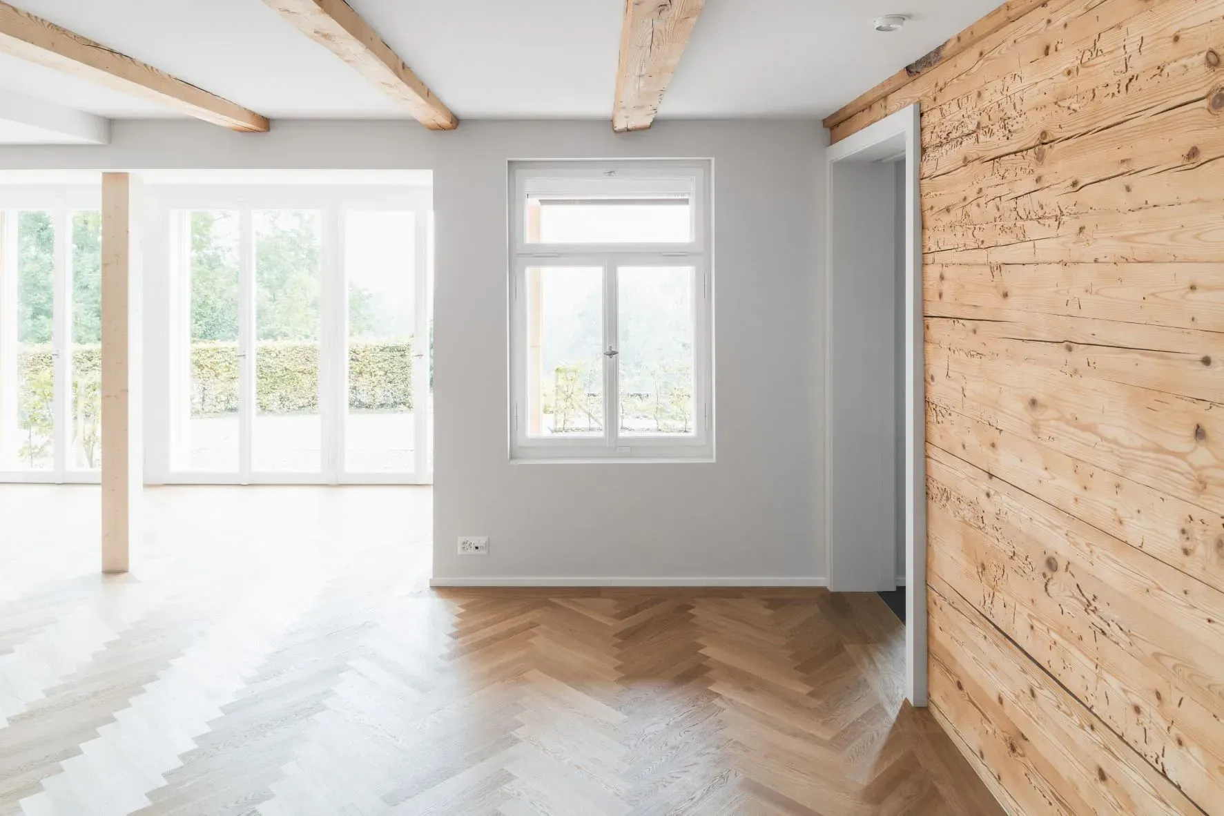 Heller, leerer Raum mit großen Fenstern, weißen Wänden, Holzbalken und Fischgrätparkett.