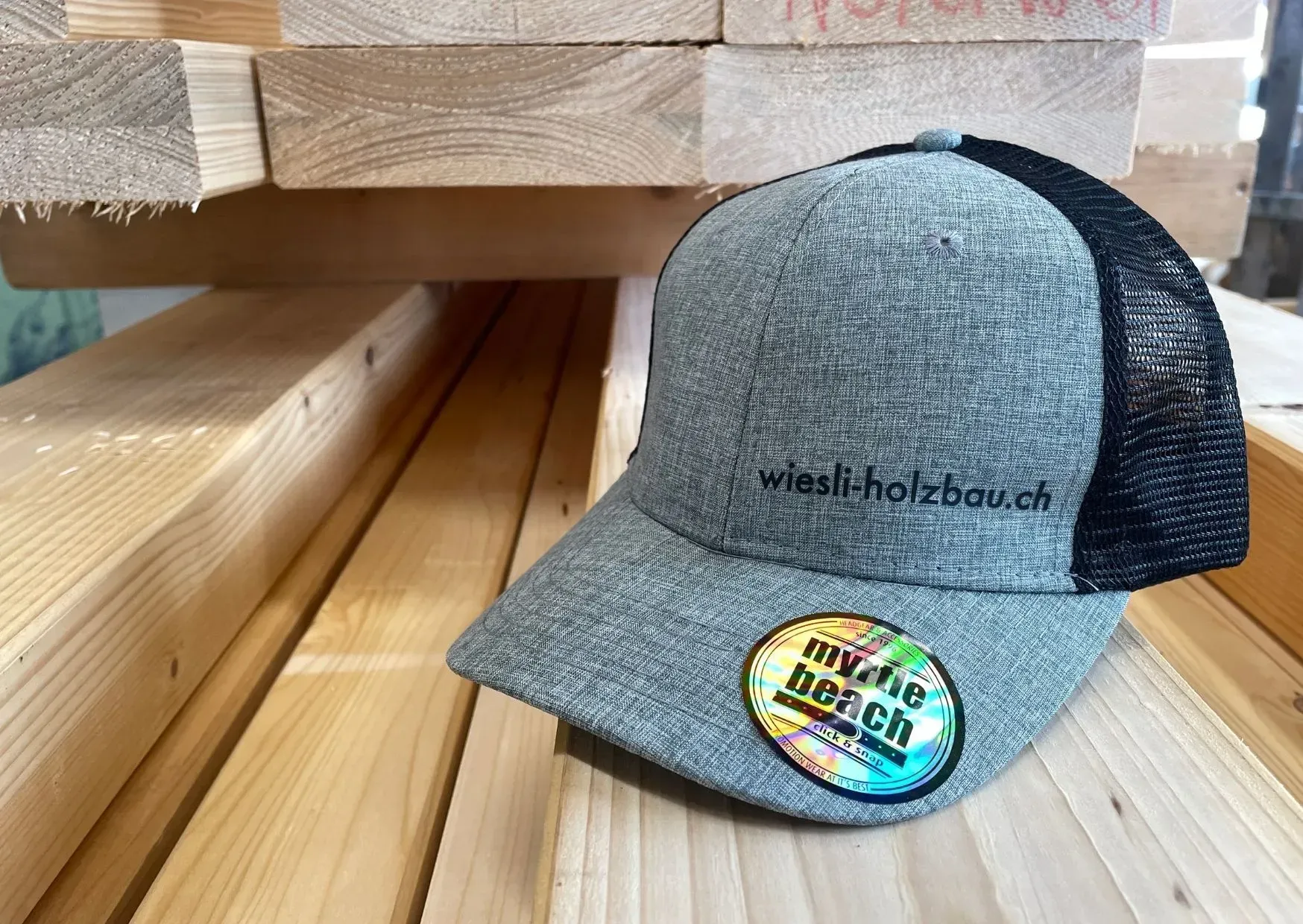 Graue und schwarze Truckerkappe aus Mesh mit Logo am Schirm, die auf gestapelten Holzbrettern ruht.