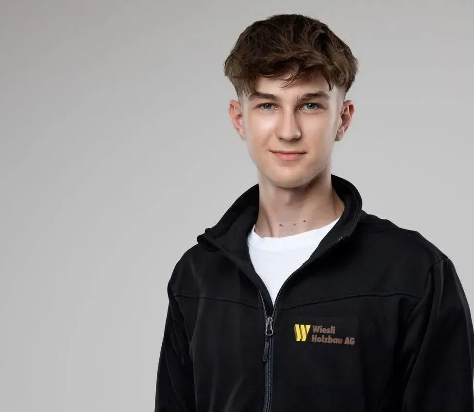 Porträt einer Person in einer schwarzen Jacke mit Logo, vor hellgrauem Hintergrund