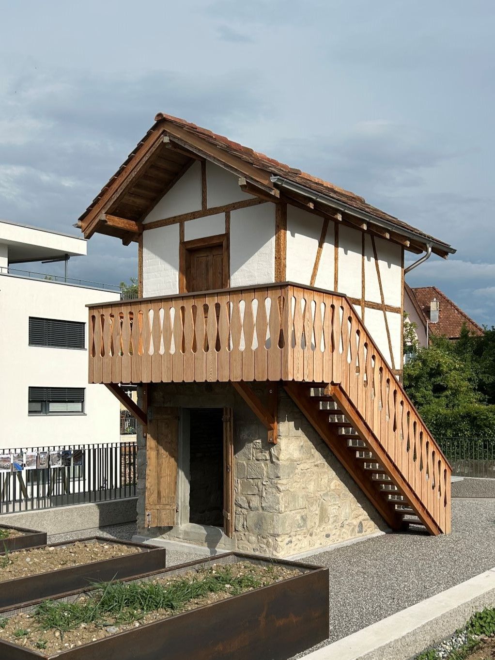 Kleines zweistöckiges Haus mit Holzbalkon und Außentreppe auf Steinsockel unter einem bewölkten Himmel