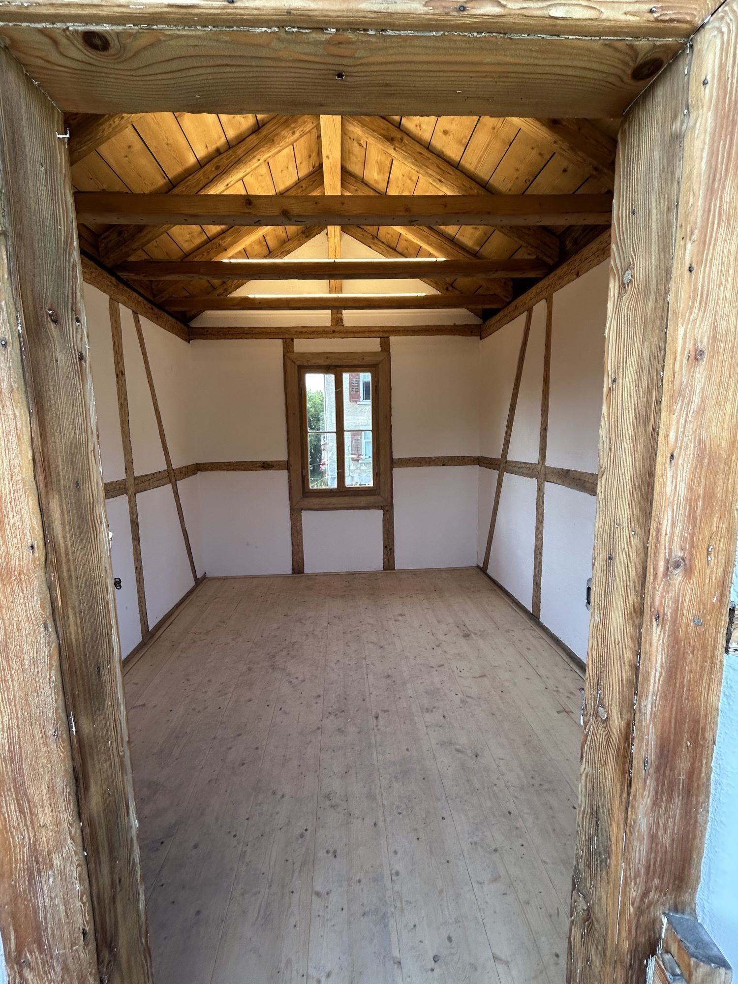 Leeres, rustikales Zimmer mit freiliegenden Holzbalken, weißen Wänden und einem kleinen Fenster am anderen Ende