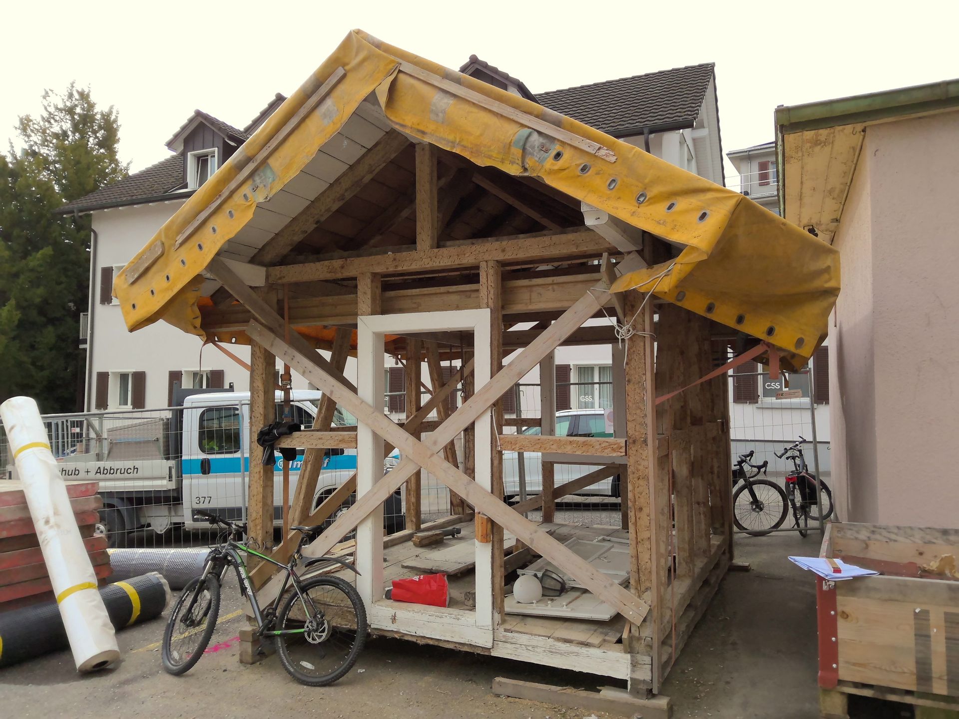 Ein kleiner, im Bau befindlicher Holzschuppen mit gelbem Planendach, daneben an der Straße lehnen Fahrräder.