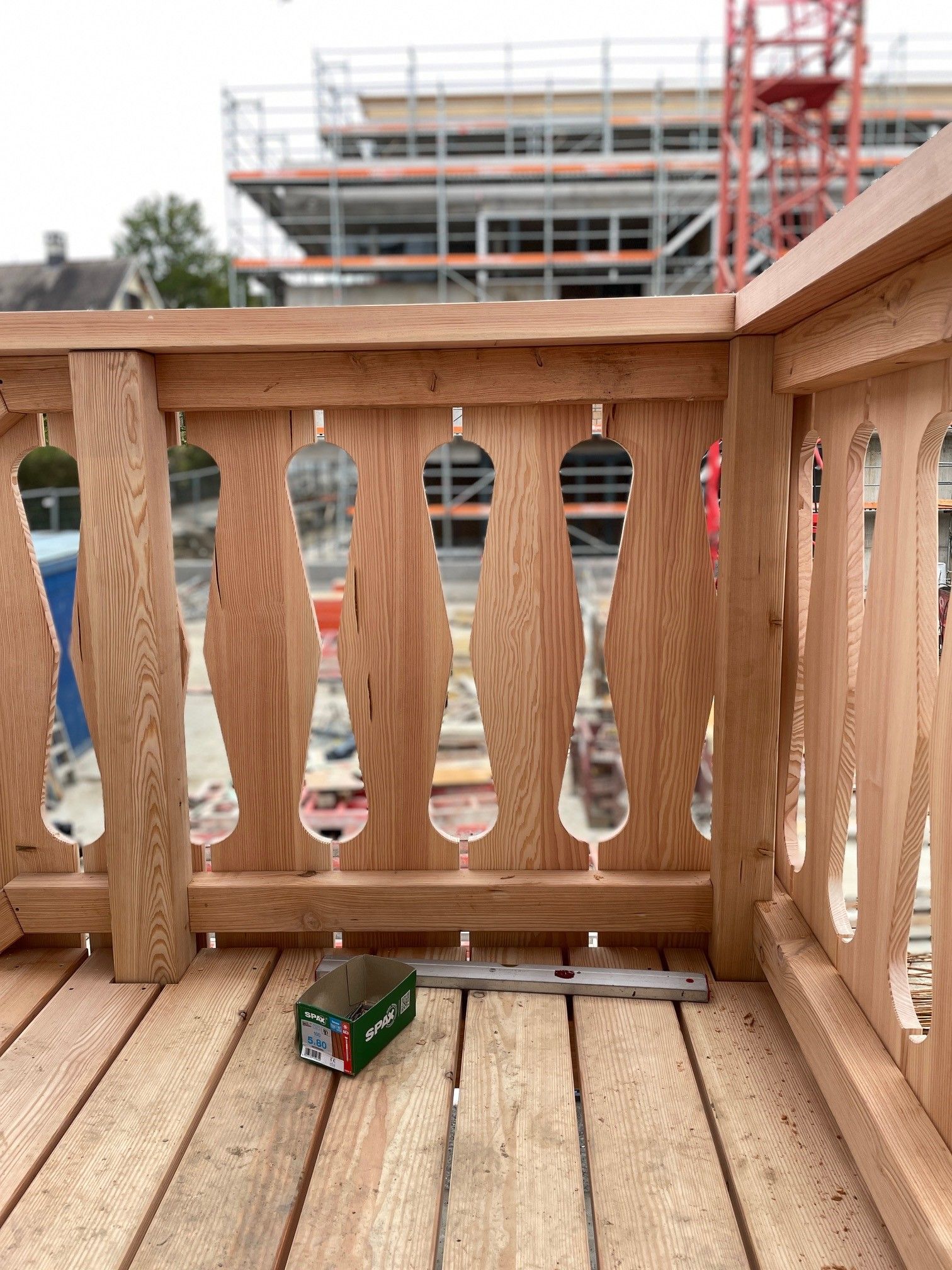 Holzbalkon mit Geländer, im Hintergrund eine Baustelle und eine kleine grüne Kiste auf dem Boden.