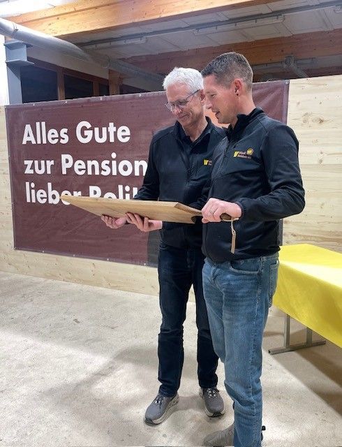 Zwei Männer in schwarzen Hemden halten eine gerahmte Gedenktafel neben einem Banner mit der Aufschrift „Alles Gute zur Pension, lieber Peter“.