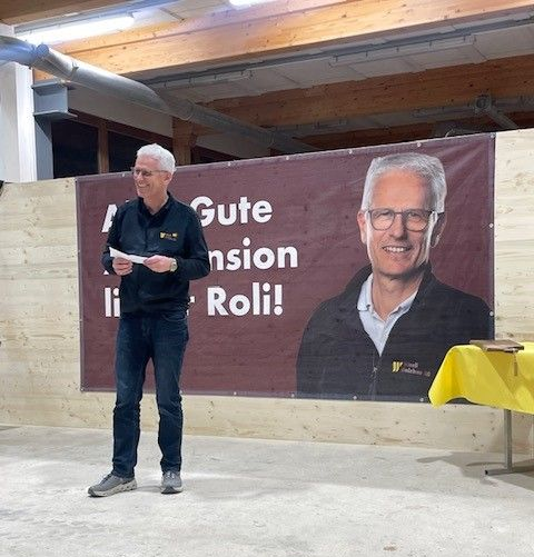 Ein Mann spricht auf einer Bühne vor einem Banner mit einem Porträt und Text und hält Papiere in einer Halle.