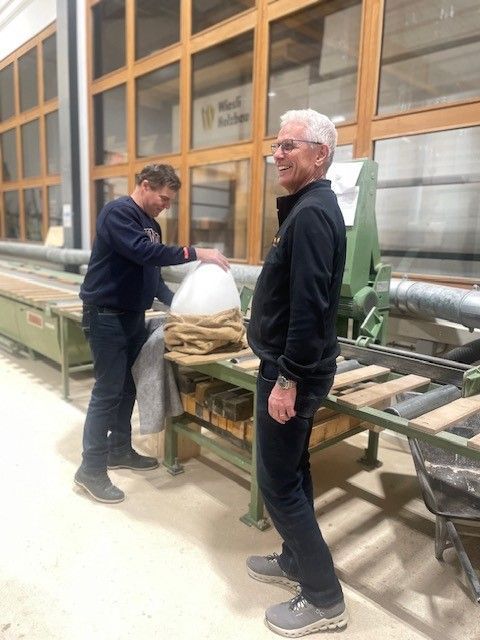 Zwei Männer stehen neben Industrieanlagen in einer Werkstatt, einer hält eine weiße Tasche in der Hand, der andere schaut in die Kamera.