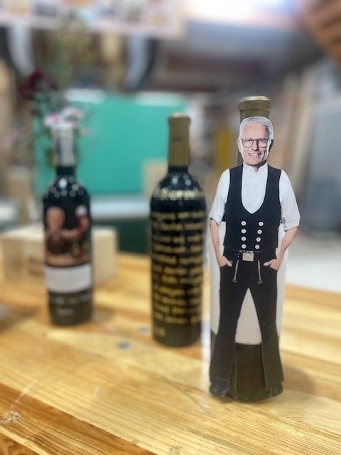 Ausgeschnittene Figur eines älteren Mannes neben zwei dunklen Flaschen auf einem Holztisch, im Hintergrund unscharf