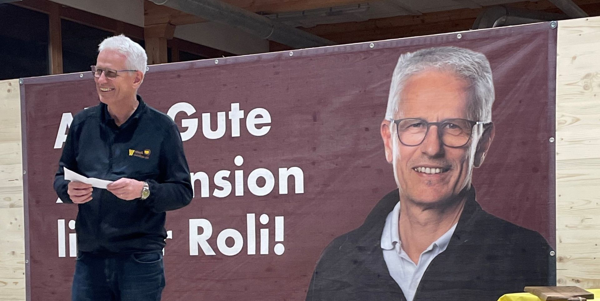 Ein Mann steht vor einem weinroten Banner mit einem großen Porträt und den Worten „Gute ... in Roli!“