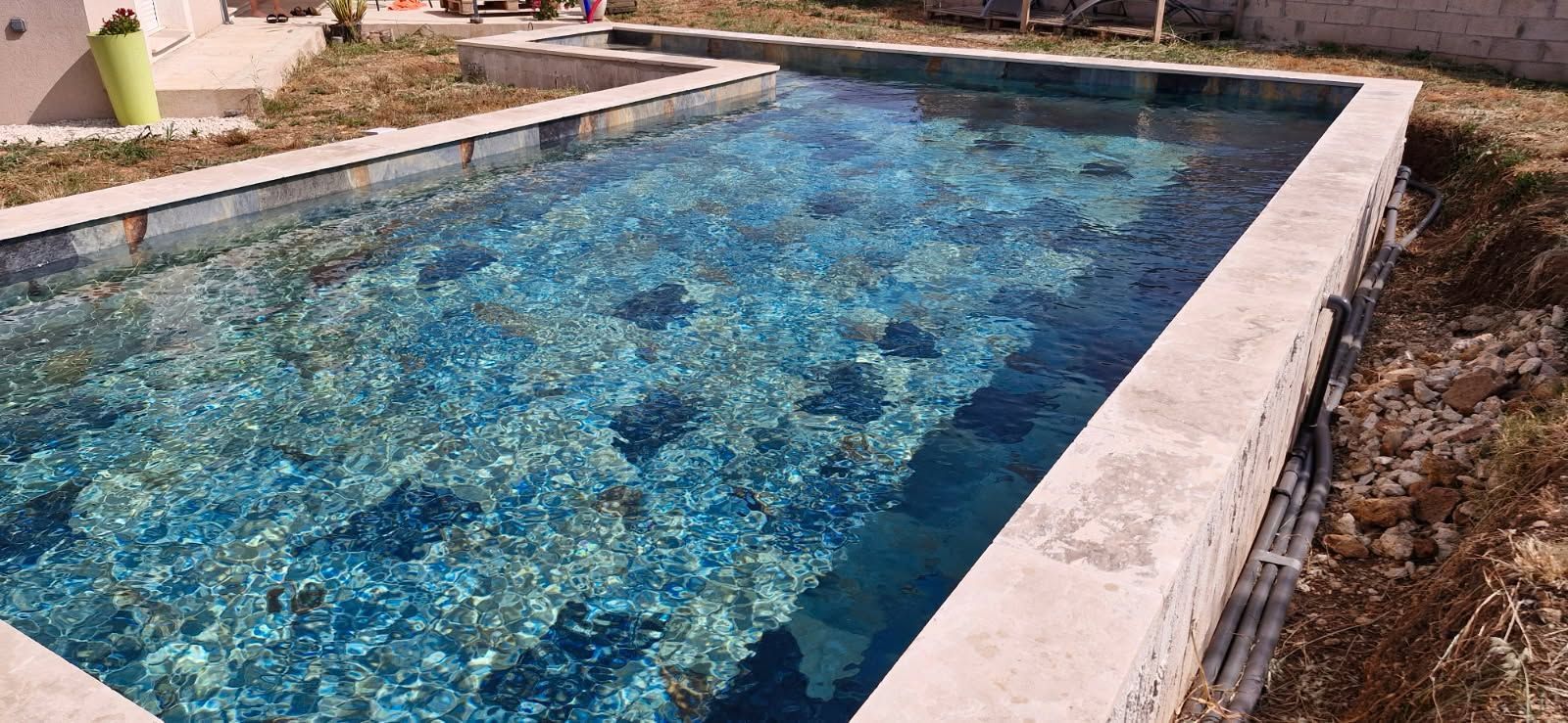 Une piscine étroite carrelée de bleu jouxte une terrasse en pierre et un aménagement paysager sec.