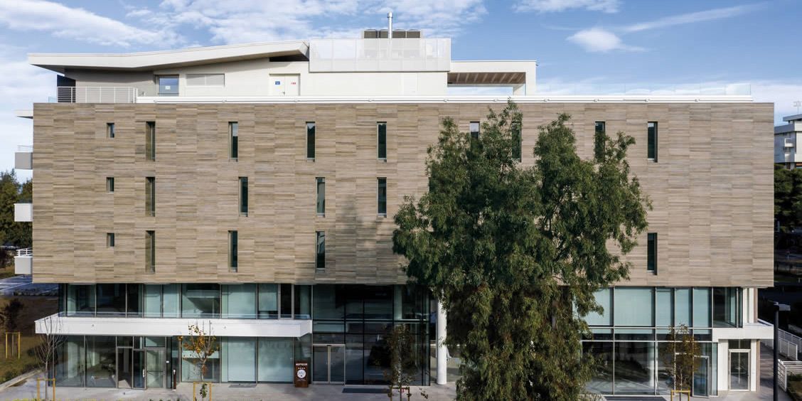 Immeuble moderne à plusieurs étages, avec une façade beige et de grandes fenêtres, partiellement masquées par un arbre.