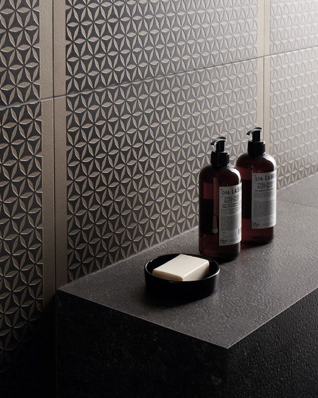 Deux distributeurs de savon et un pain de savon sur un comptoir sombre, à côté d'un mur carrelé à motifs.