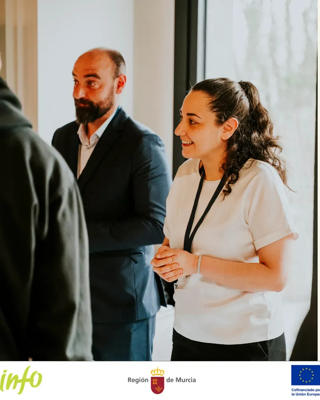 Dos personas vestidas de negocios sonríen y conversan en un evento en interiores con ventanas luminosas.
