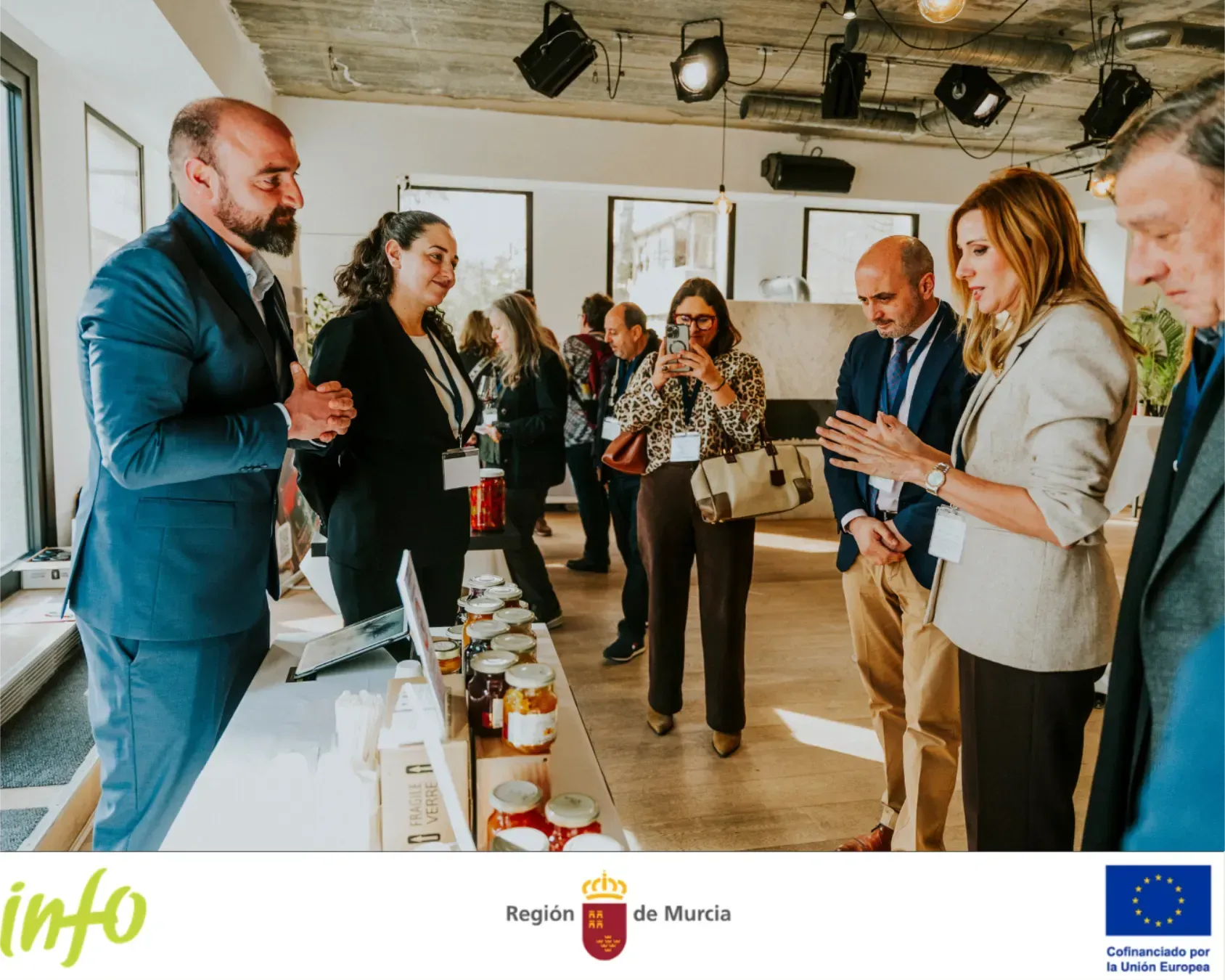 La gente socializa en una luminosa recepción interior, charlando alrededor de una mesa con bebidas y aperitivos.