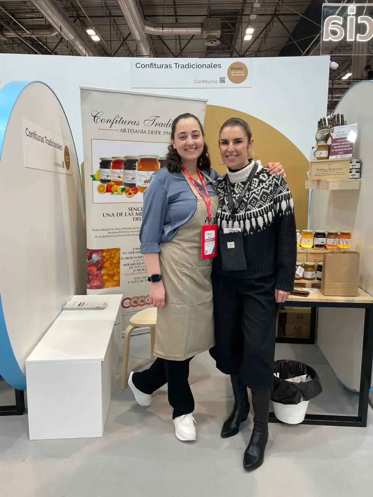 Dos mujeres posando en un stand de feria comercial con exhibidores de productos y estantes de empaque.