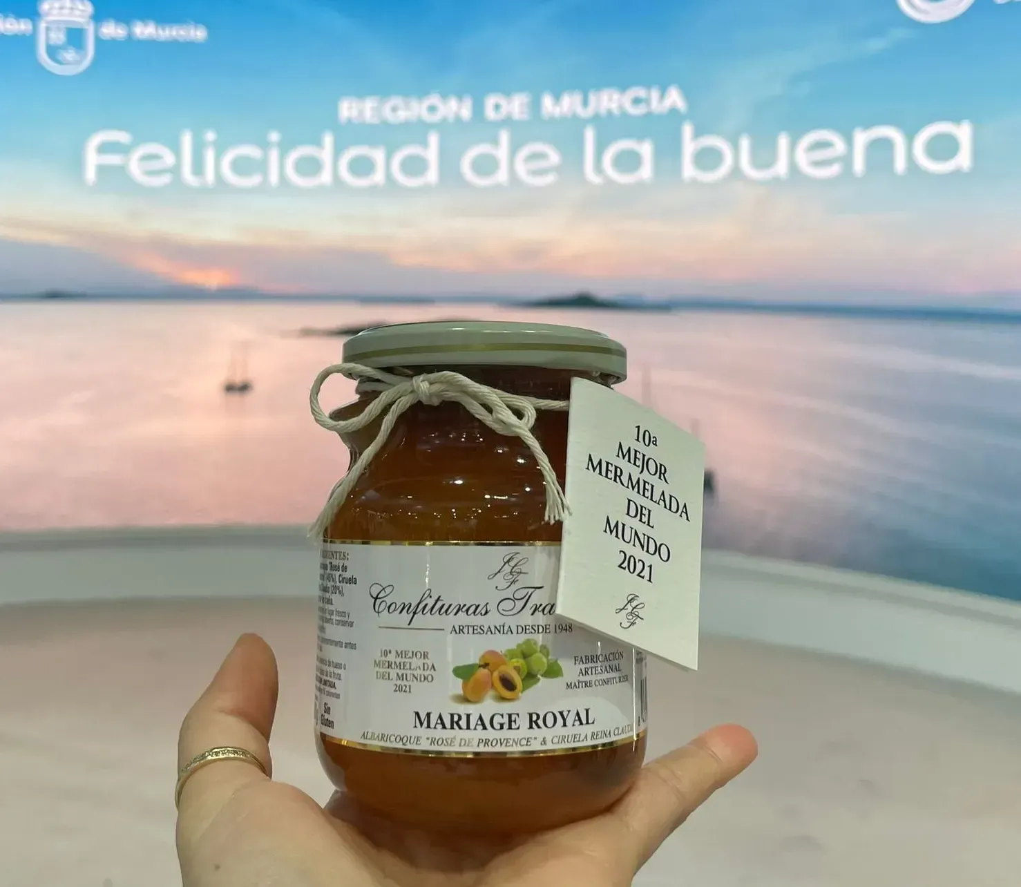 Una mano sostiene un tarro de miel frente a un cartel de “Región de Murcia” al atardecer junto al mar.