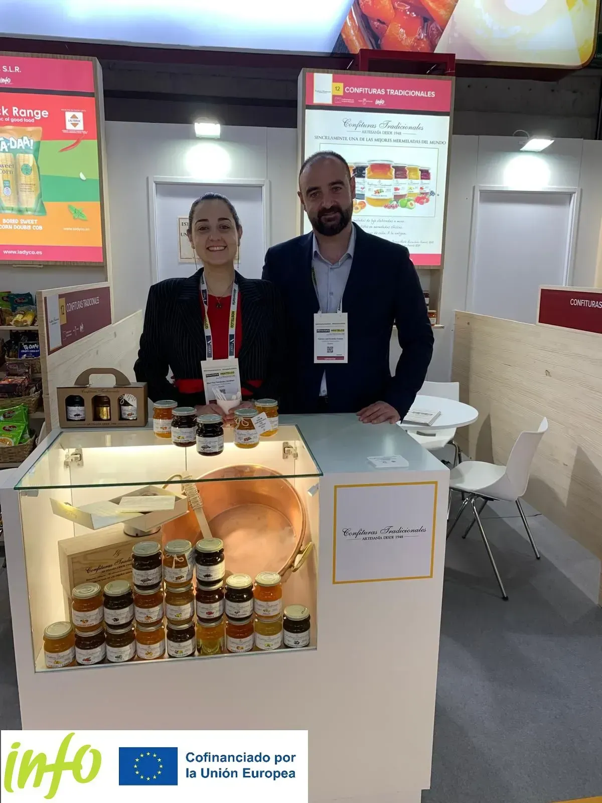 Dos personas en un puesto comercial exhibiendo frascos de productos sobre una mesa iluminada.