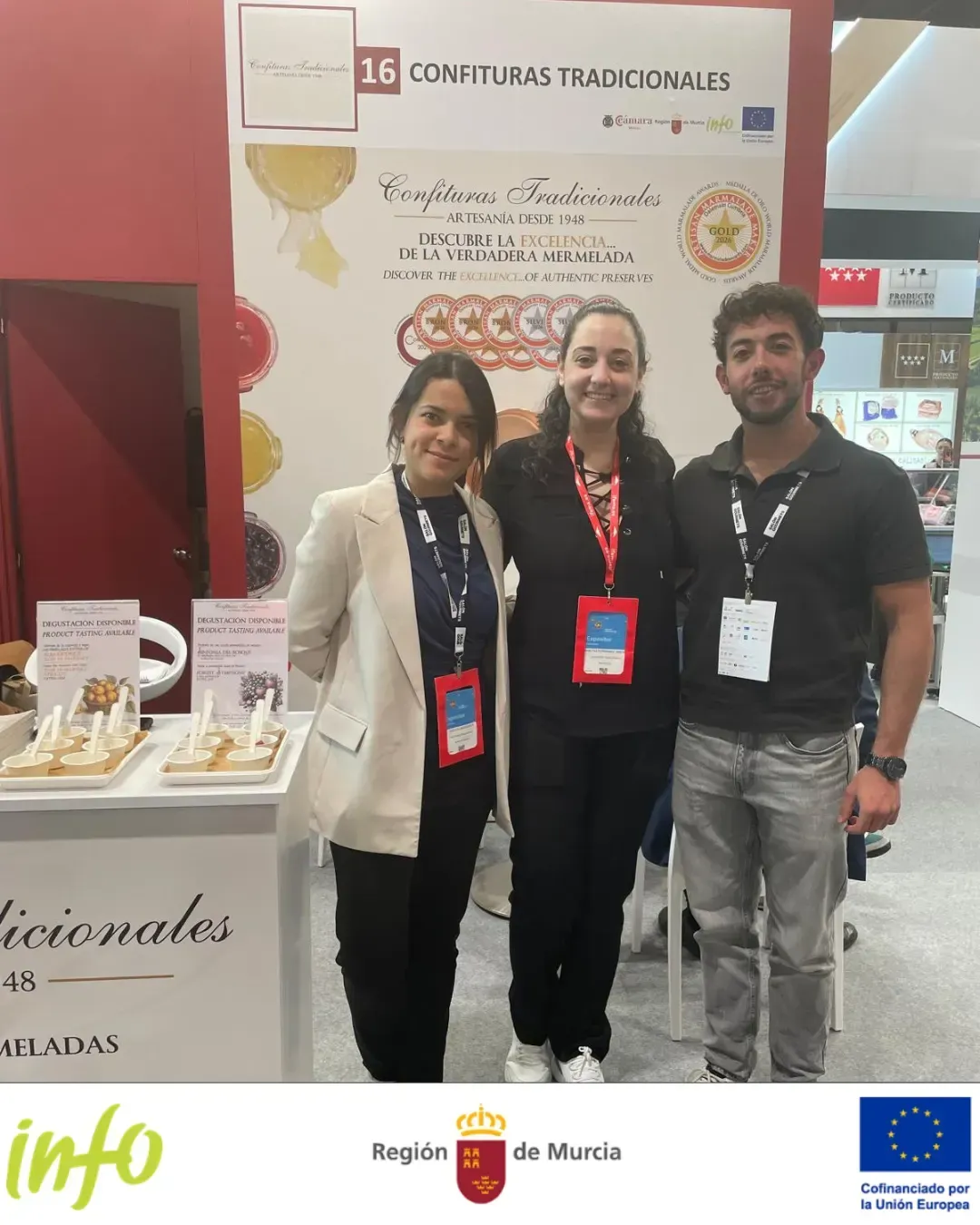 Tres personas posando en un stand de feria con un fondo de exhibición beige y detalles en rojo.