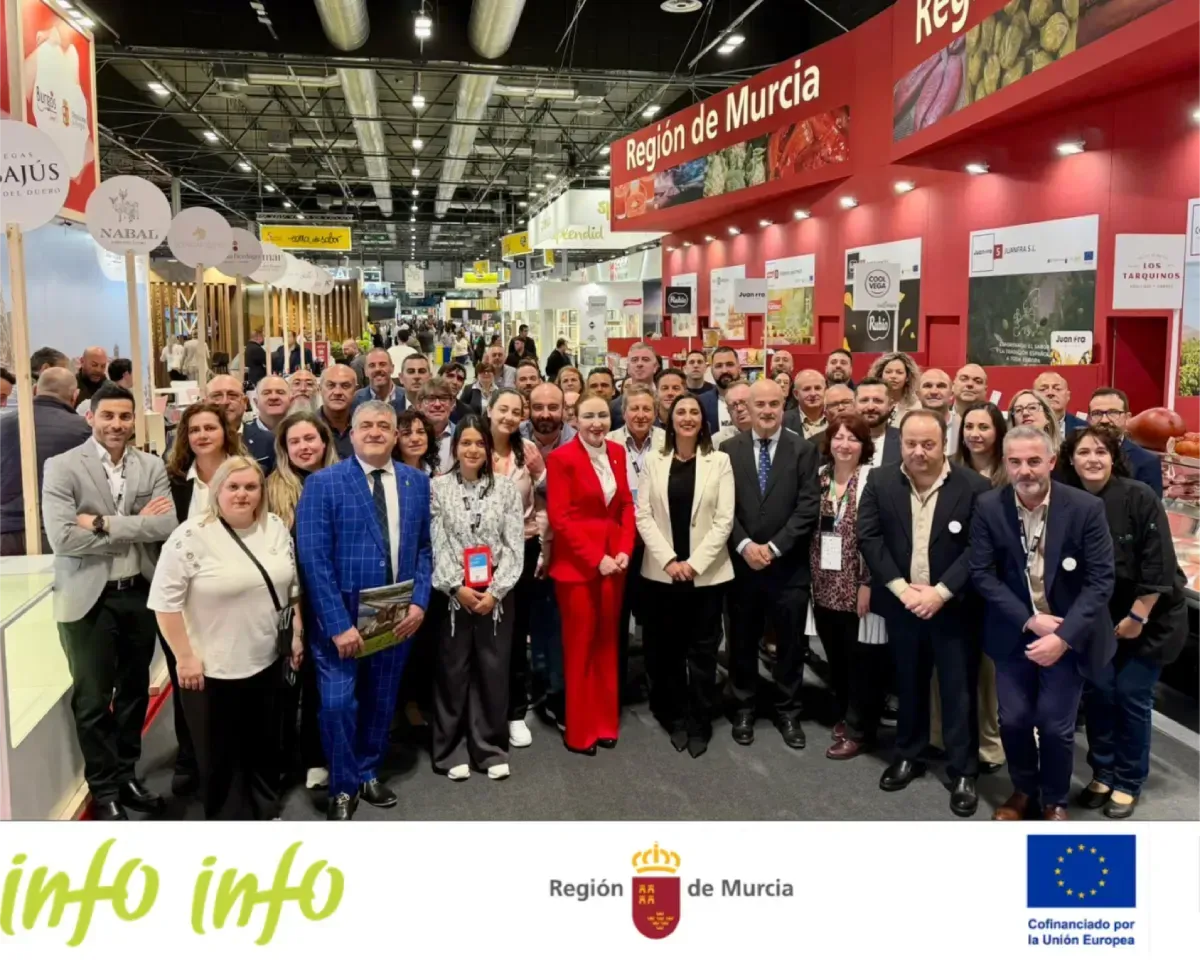 Grupo numeroso posando en un stand de feria con letreros rojos de “Región de Murcia” y mostradores con la marca.
