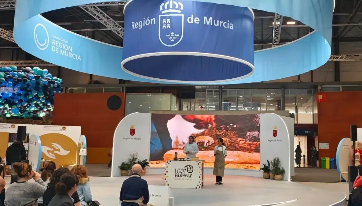 Stand de turismo de Murcia en una feria comercial, con carteles azules colgantes, pantallas de escenario