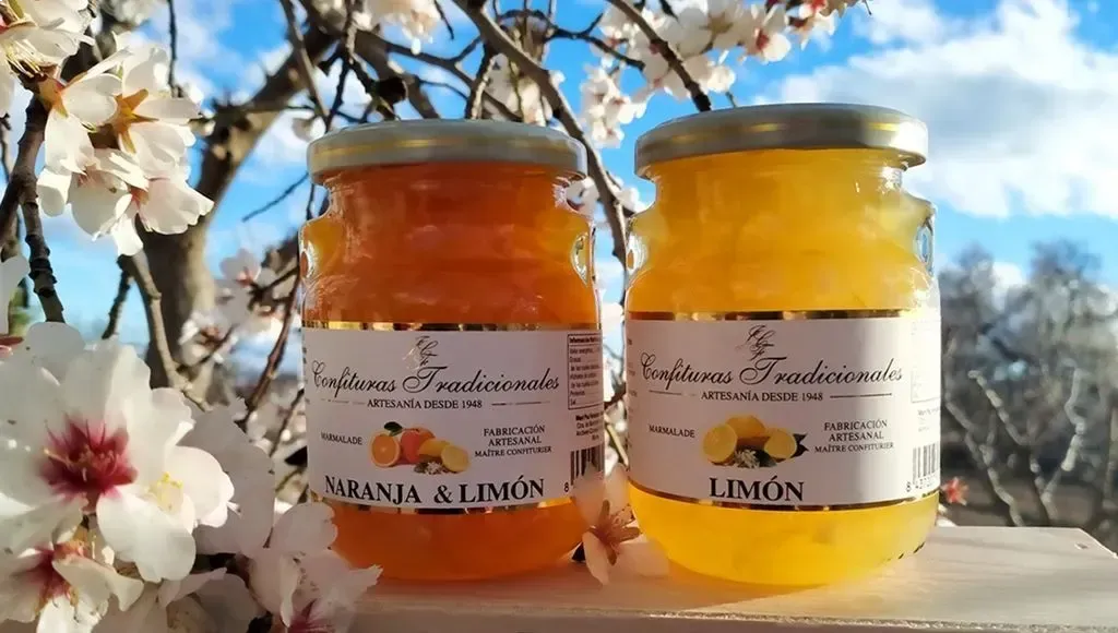 Dos tarros de miel, uno naranja y otro amarillo, sobre una repisa con cerezos en flor al fondo.