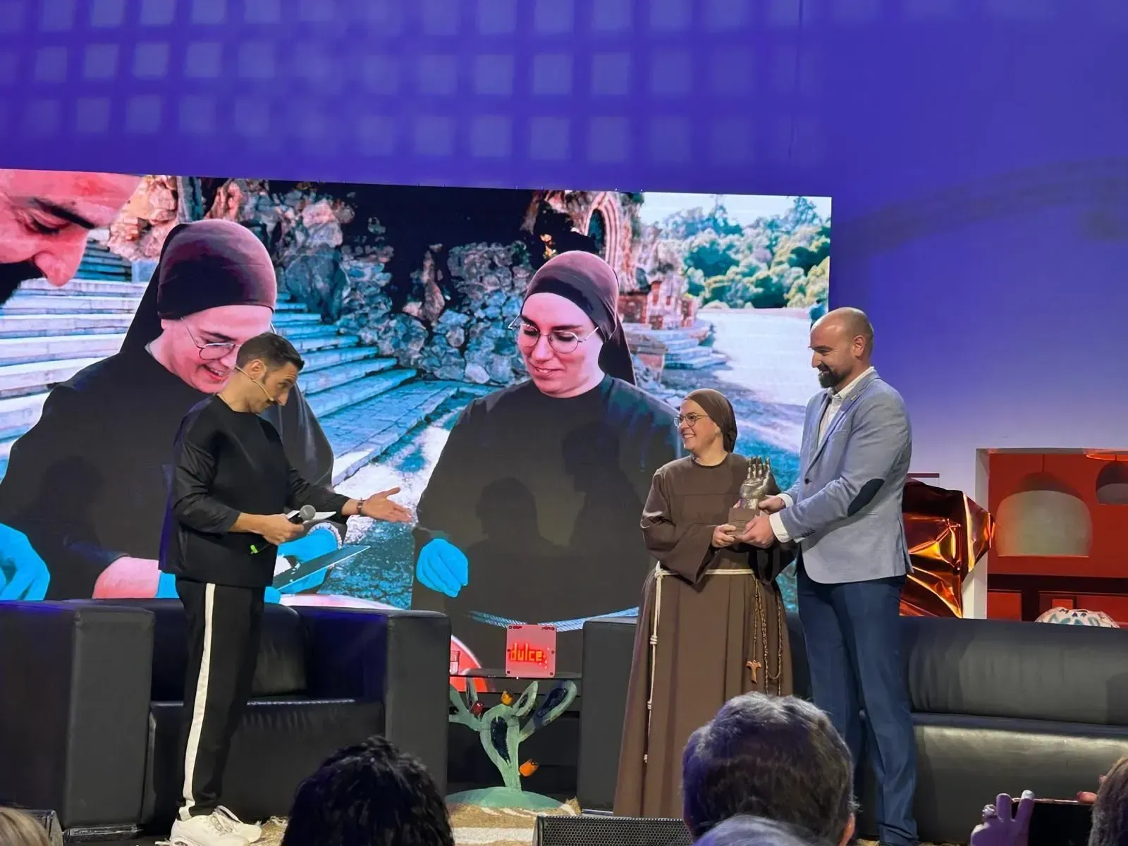 Panel en el escenario con un gran telón de fondo para fotos; tres presentadores están de pie cerca de un sofá y una pantalla.