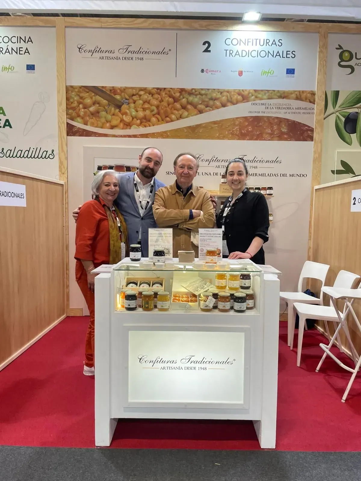 Cuatro personas se encuentran detrás de un stand de exhibición con frascos y bandejas en una feria comercial.
