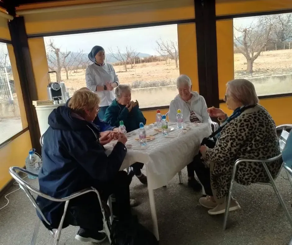 Varias personas sentadas alrededor de una mesa en una habitación soleada, con una persona de pie cerca.