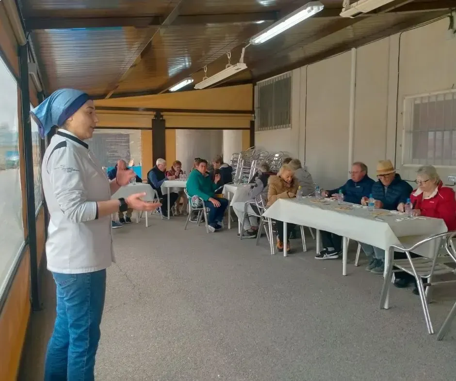 Presentador dirigiéndose a un público sentado en un salón cubierto con mesas largas y sillas blancas.