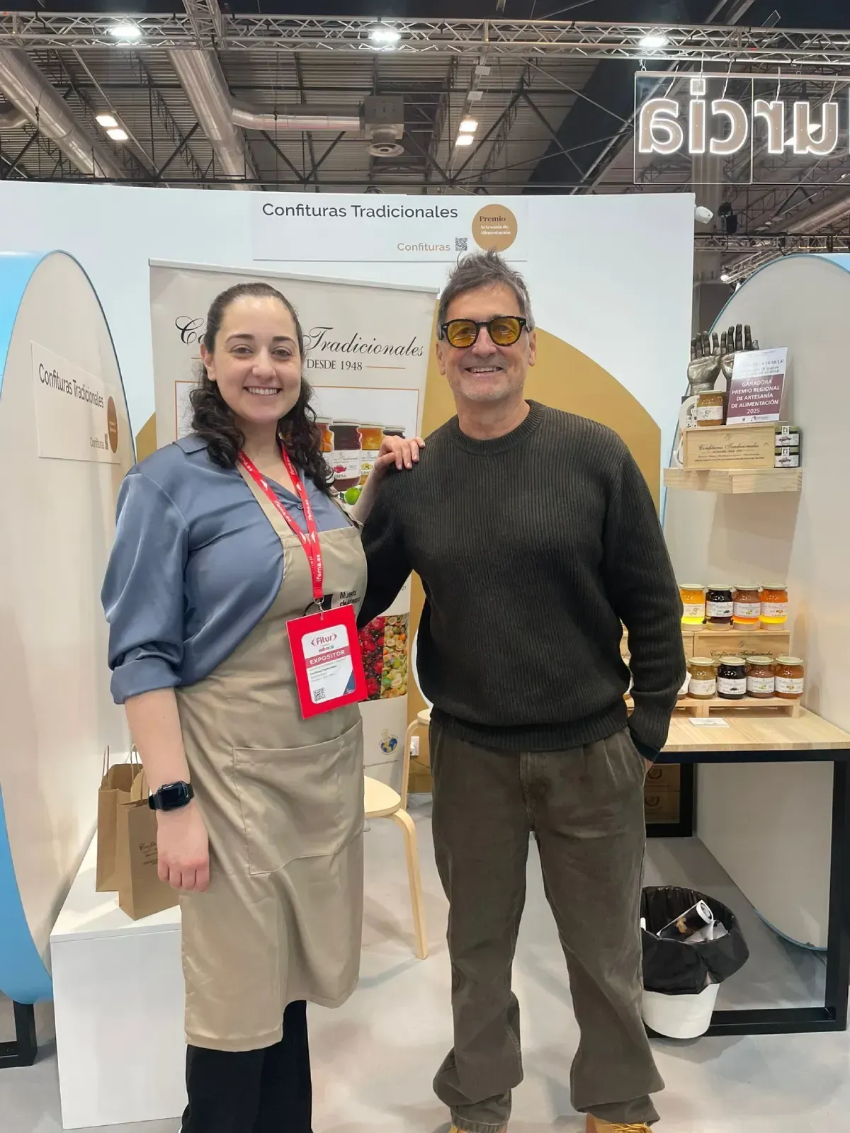 Dos personas sonriendo en un stand de una feria comercial, de pie junto a tablas de surf y expositores de productos.