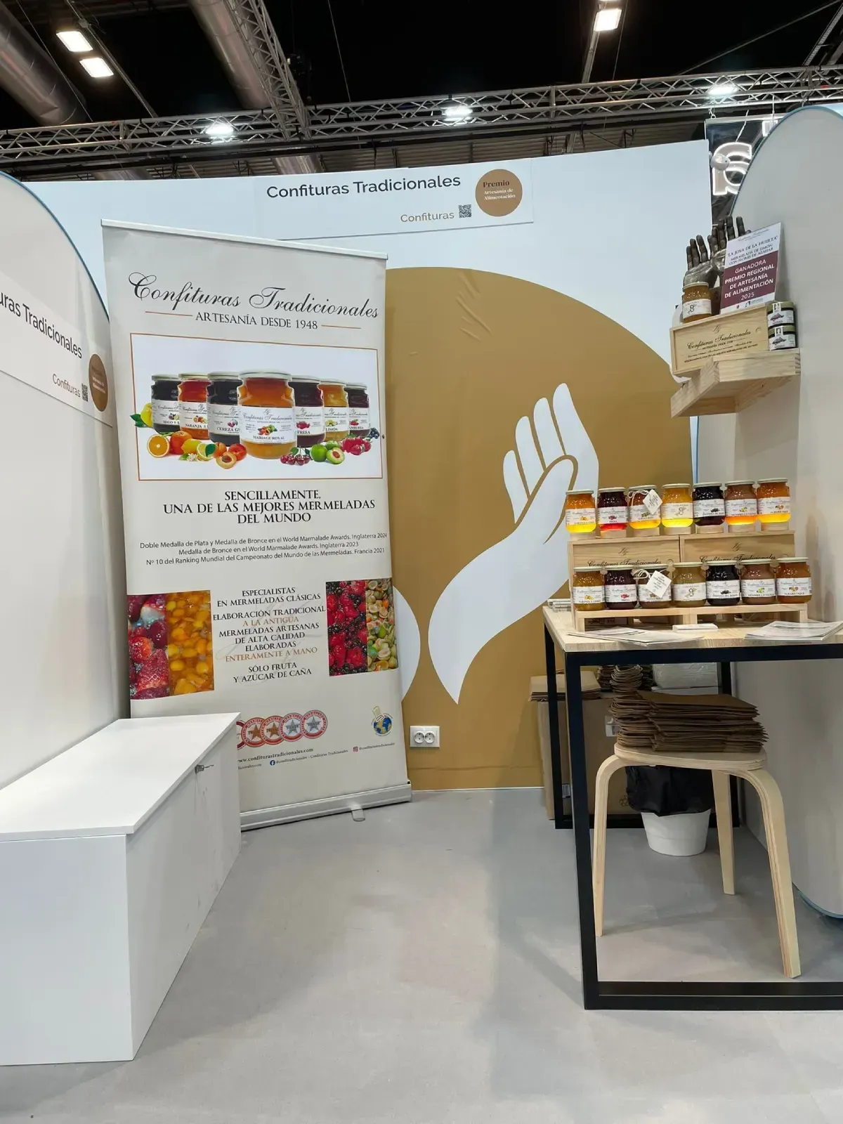 Stand de feria comercial con pancarta blanca, pared color canela, estantes para refrigerios y una pequeña mesa de exhibición.