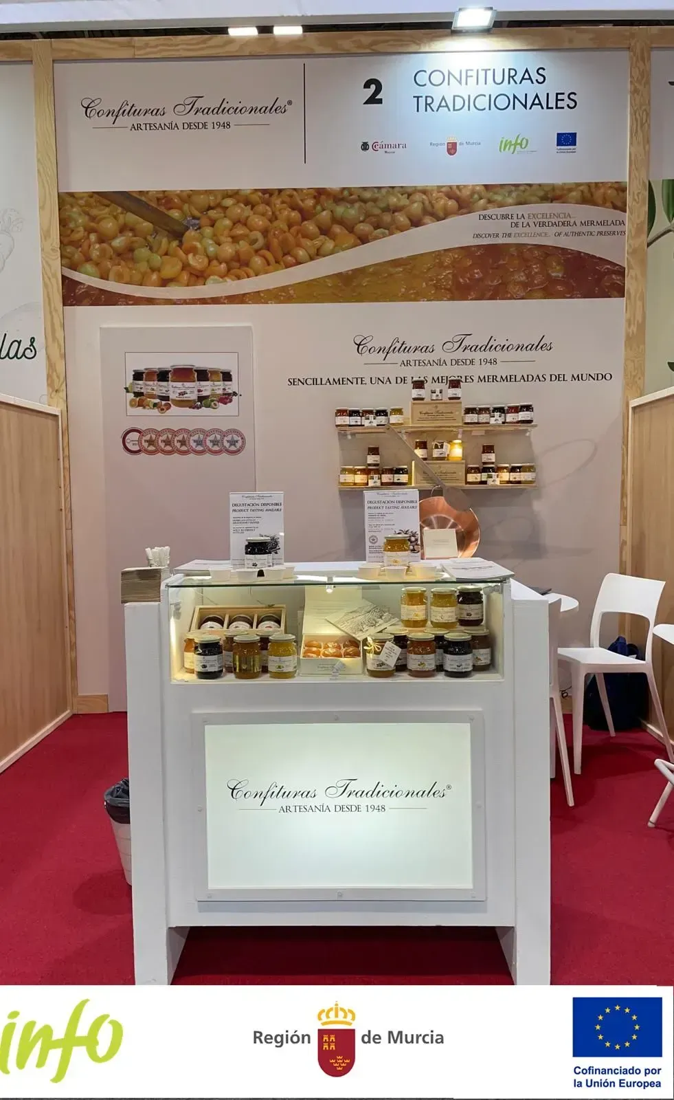 Stand de feria comercial con mostrador blanco, exhibición de alimentos y telón de fondo con pancarta en un pabellón