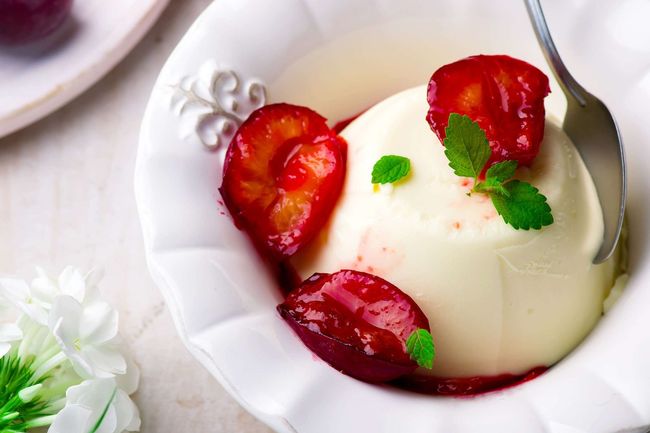 Postre de panna cotta en recipiente blanco, cubierto con rodajas de ciruela roja y hojas de menta.