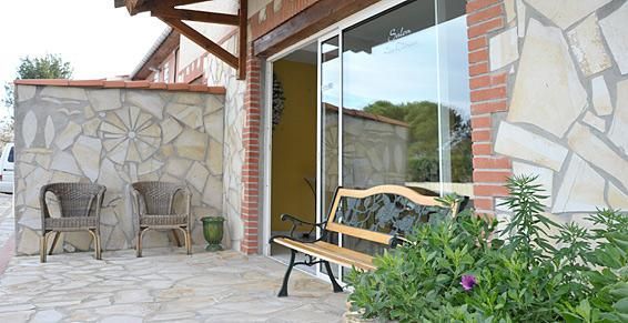 Espace salon extérieur avec murs en pierre, chaises en osier, banc en bois et grande porte vitrée.