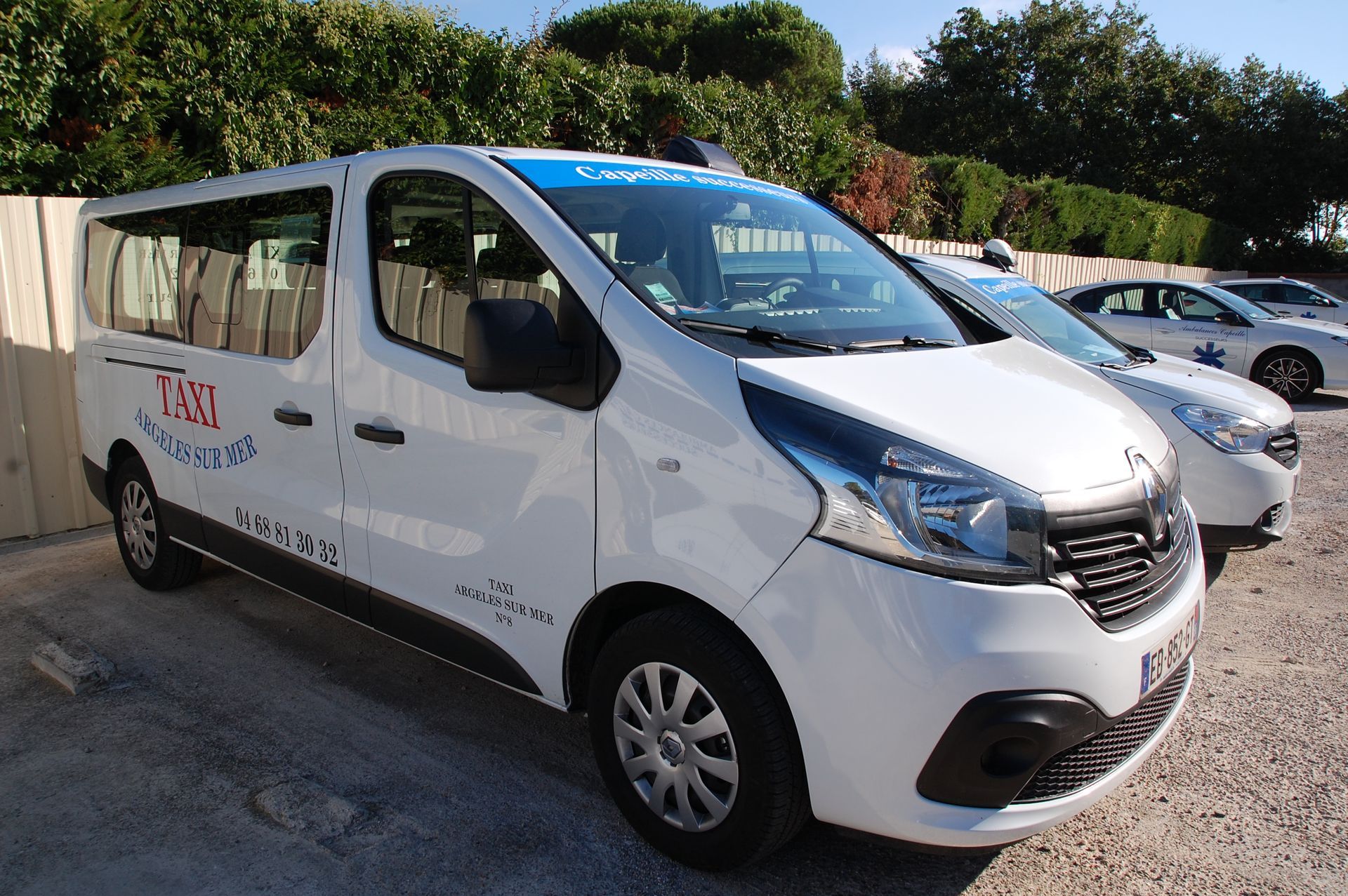 Une camionnette blanche de transport de passagers, portant le logo de l'entreprise, est garée à l'extérieur, à côté d'une voiture, sous un ciel bleu.