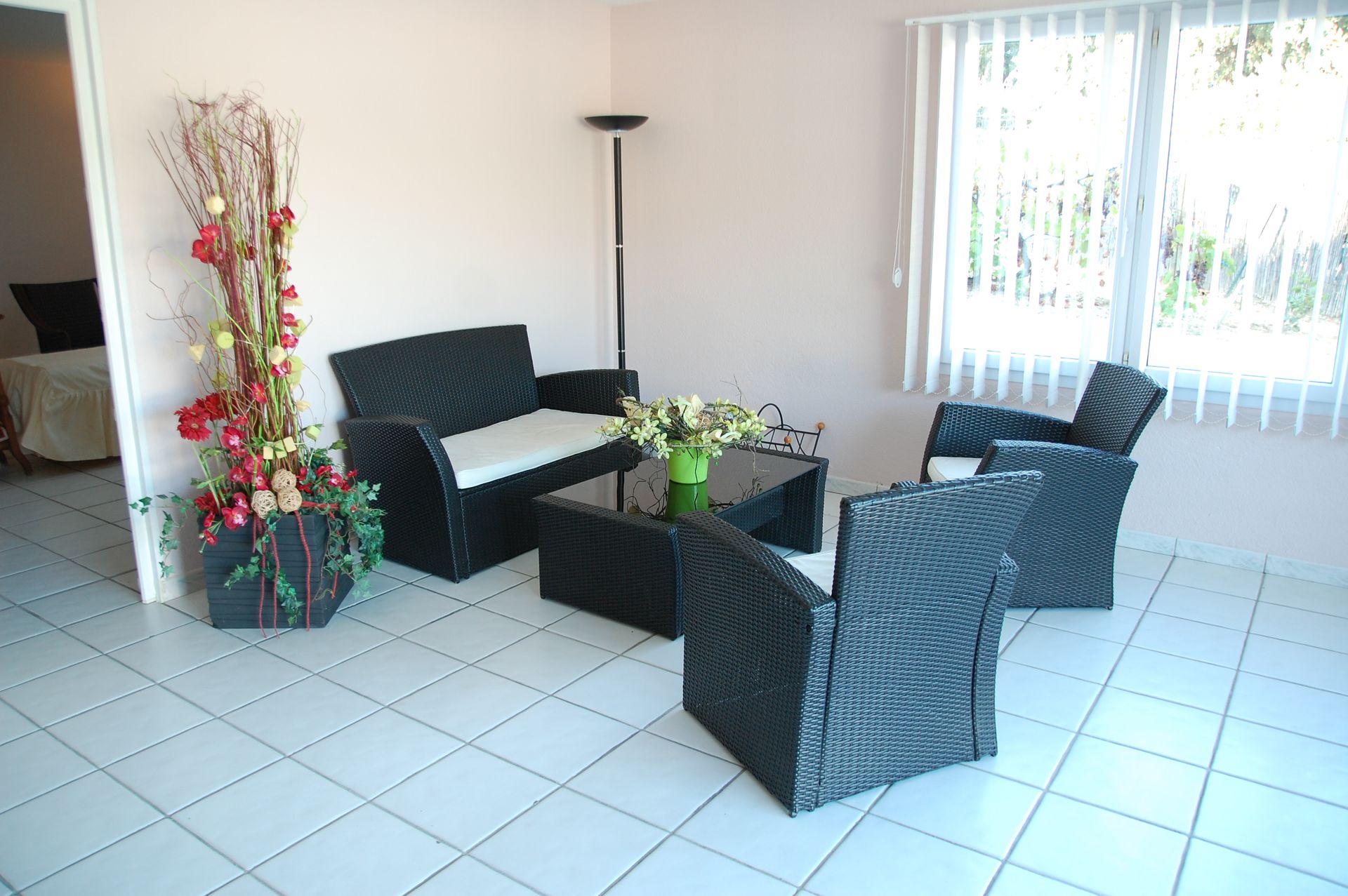 Salon avec meubles en osier noir, décoration florale et sol carrelé blanc.