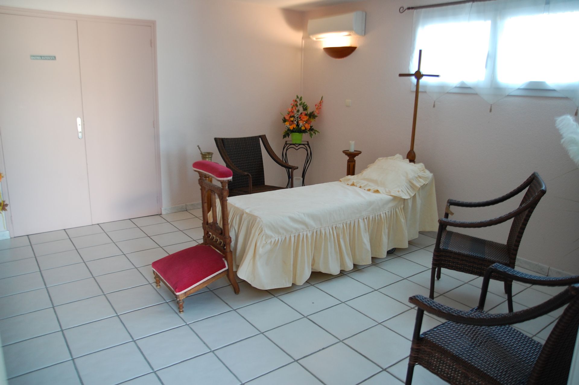 Chambre funéraire avec un lit, des chaises et une croix. Murs roses et sol carrelé.