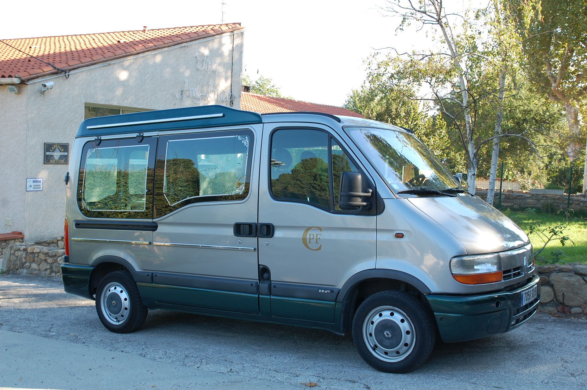 Une camionnette argentée et verte est garée à côté d'un bâtiment au toit de tuiles rouges, dans un cadre naturel.
