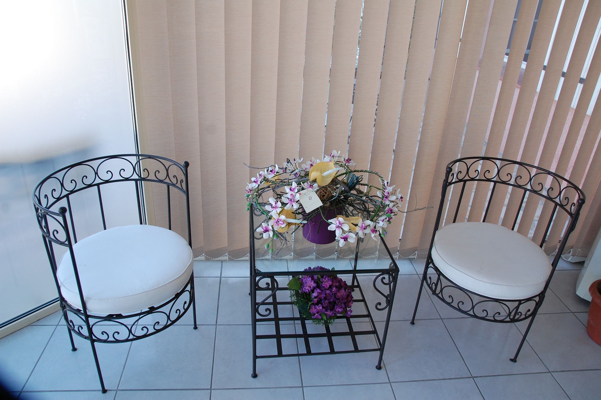 Coin salon sur le balcon avec chaises en fer forgé, table avec fleurs et stores.