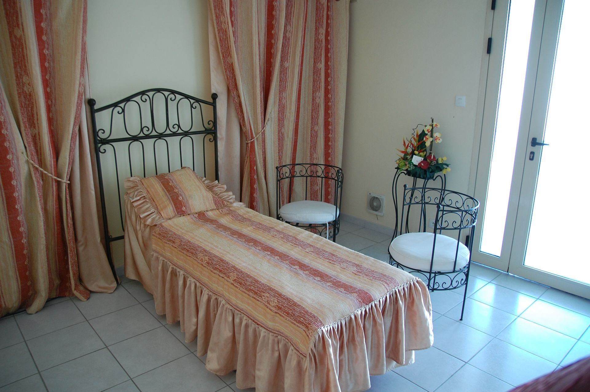 Chambre avec lit en fer forgé, deux chaises et rideaux à motifs. Ambiance lumineuse et aérée.