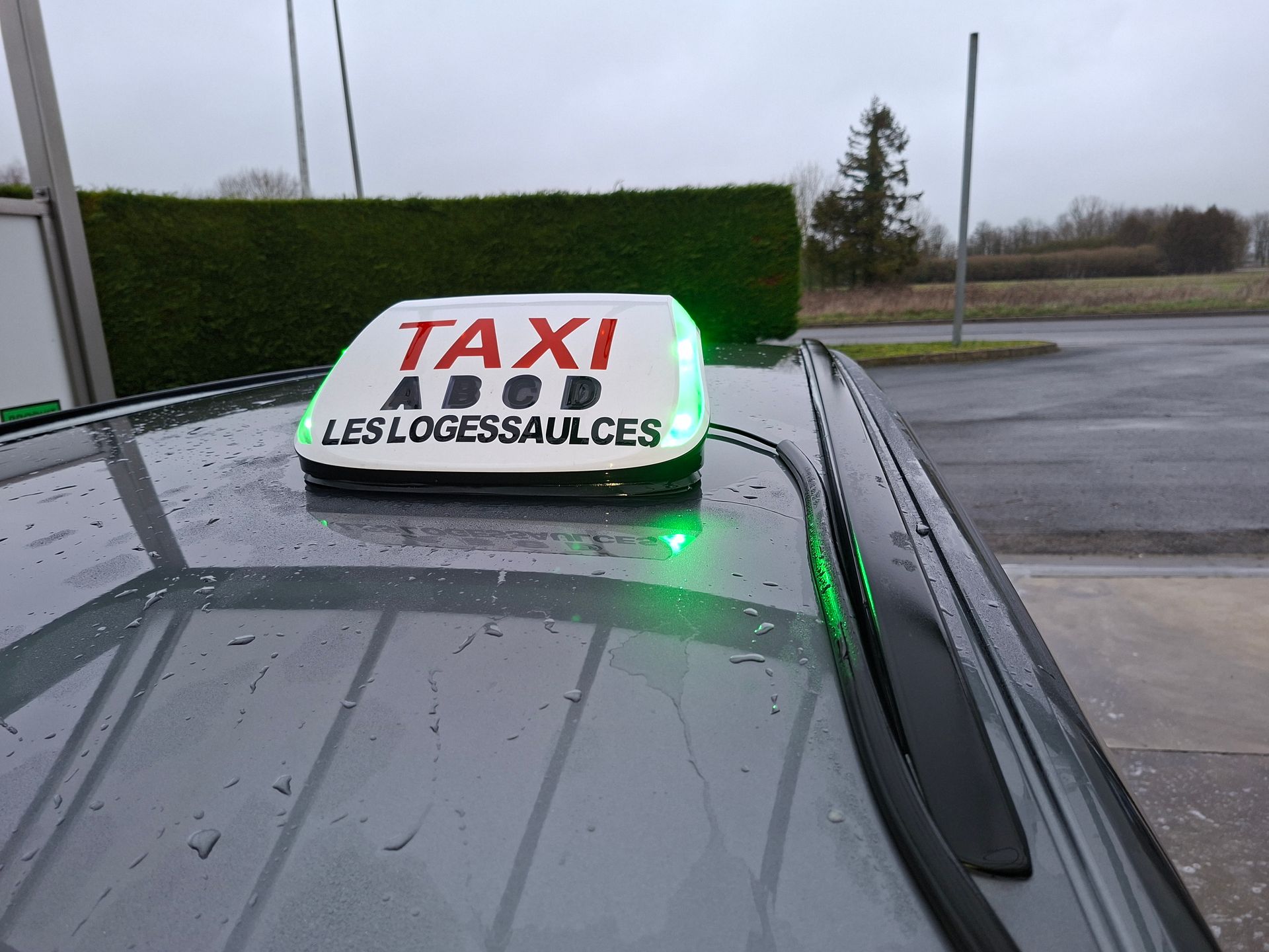 Un panneau blanc sur le toit d'un taxi, portant l'inscription TAXI ABCD LES LOGESSAULCES, avec des lumières vertes allumées, sur le toit gris d'une voiture.