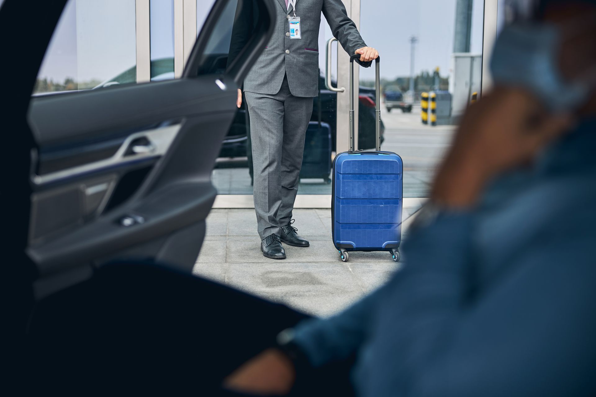 Une personne sortant d'une voiture, vêtue d'un costume et portant des bagages, près d'une entrée d'aéroport.
