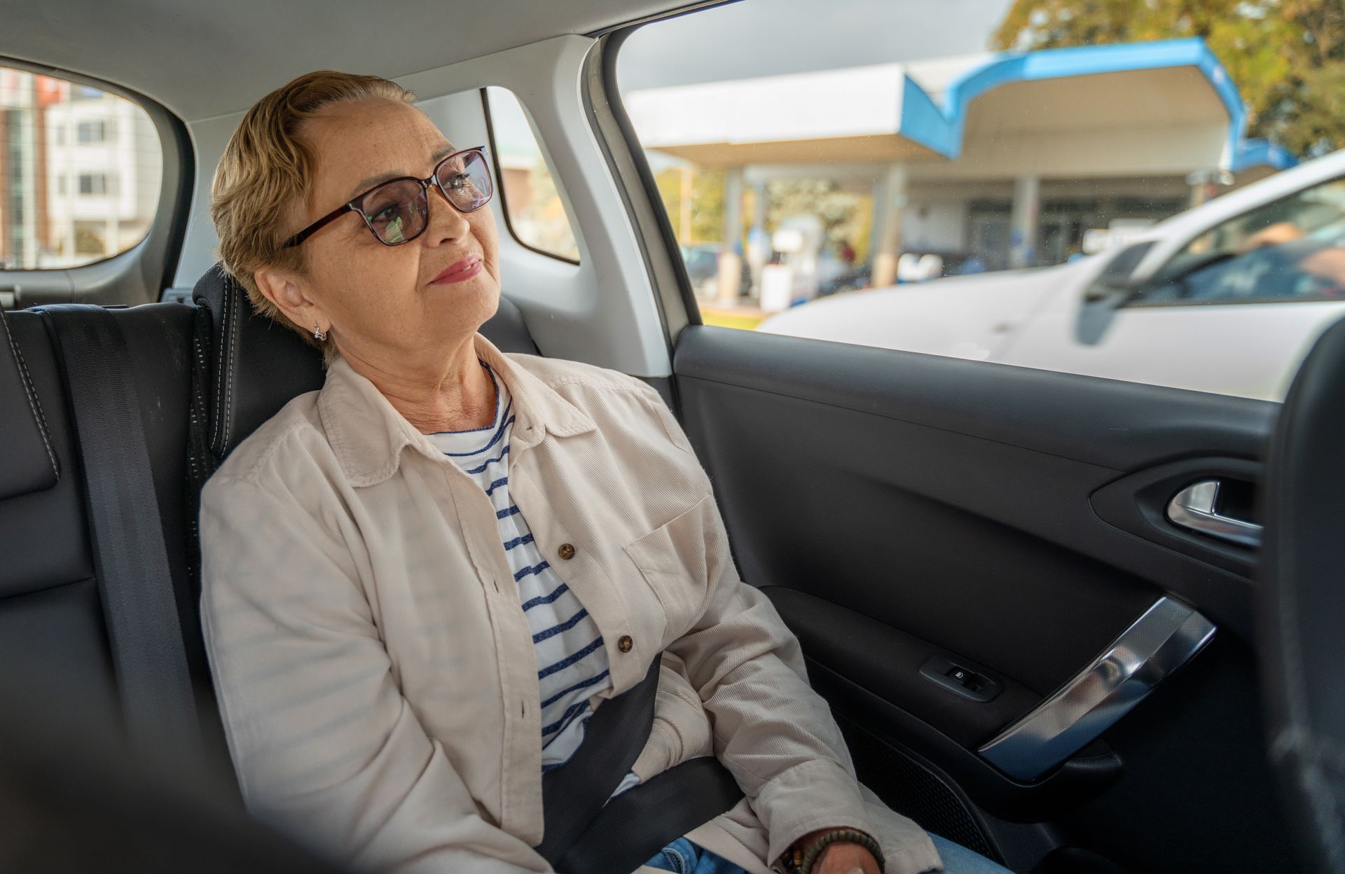 Une femme dans une voiture, portant des lunettes de soleil et une ceinture de sécurité, regarde par la fenêtre ; une station-service est visible.